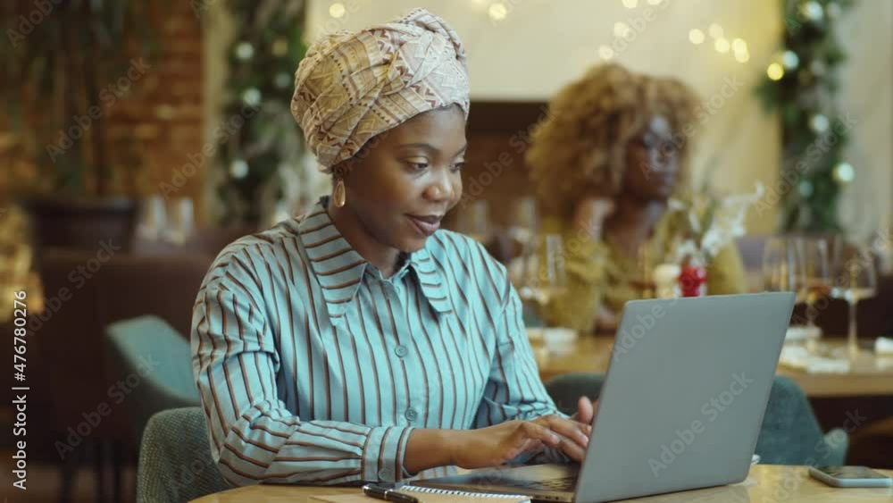 Beautiful African American woman sitting at table in cafe and working remotely on laptop Beautiful African American woman sitting at table in cafe and working remotely on laptop