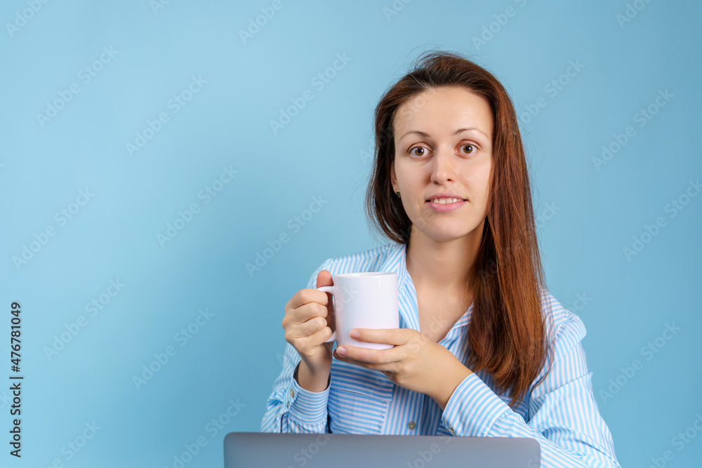 Portrait of a young adult girl with a white mug in her hands and a laptop. Blue background with copyspace Portrait of a young adult girl with a white mug in her hands and a laptop. Blue background with copyspace