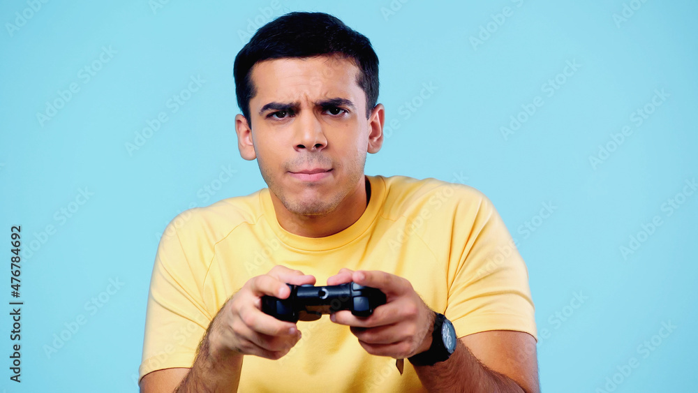KYIV, UKRAINE - DECEMBER 22, 2021: worried young man holding joystick and playing video game isolated on blue. KYIV, UKRAINE - DECEMBER 22, 2021: worried young man holding joystick and playing video game isolated on blue.