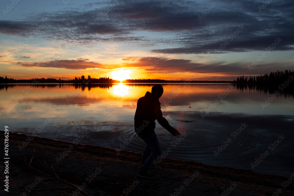 silhouette of Young Filipino man skipping rocks on lake during colourful sunset