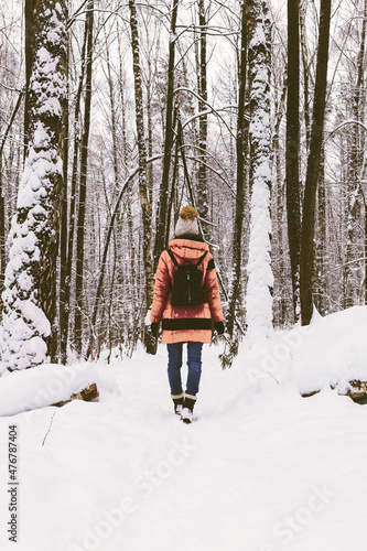 Winter forest and woman walking in snow nature. Rear view of girl in Calming Coral color down jacket and warm hat strolls in woods