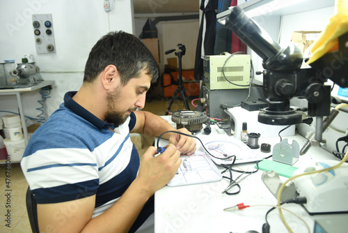 A young man at a desk in a workshop laboratory repairing a chip with an electric soldering iron