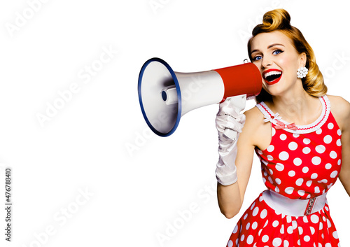 Beauty blond haired woman holding megaphone, shout something. Girl in red pin up style dress in polka dot, isolated over white background. Pinup model in retro and vintage studio concept.