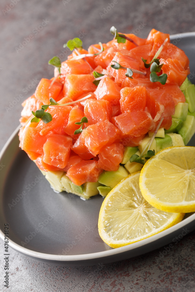 Salmon tartare on top on slices of avocado with lemon and microgreen close-up in a plate on a concrete table. Vertical