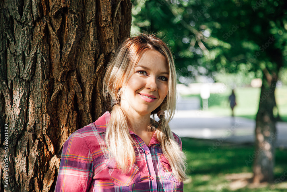 Close-up portrait of adult shining blonde woman with sun on face and with pigtails looking into camera leaning on oak in park wearing checkered shirt in daytime. Adorable background full of green. Close-up portrait of adult shining blonde woman with sun on face and with pigtails looking into camera leaning on oak in park wearing checkered shirt in daytime. Adorable background full of green.