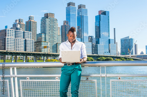 Dressing in a white shirt and green pants, a young black guy with mohawk hair is standing in the front of high buildings by the water, working on a computer..