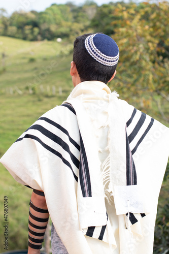 Vertical photo. Jewish man praying with talit, kippah and tefillin in nature with beautiful green meadow in the background. photo