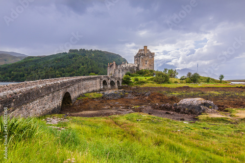 Eilean Donan Castle in summer with green grass and a stone wall in the foreground. Stone bridge over a water access. View of the fortress at low water with dark clouds and hills with trees