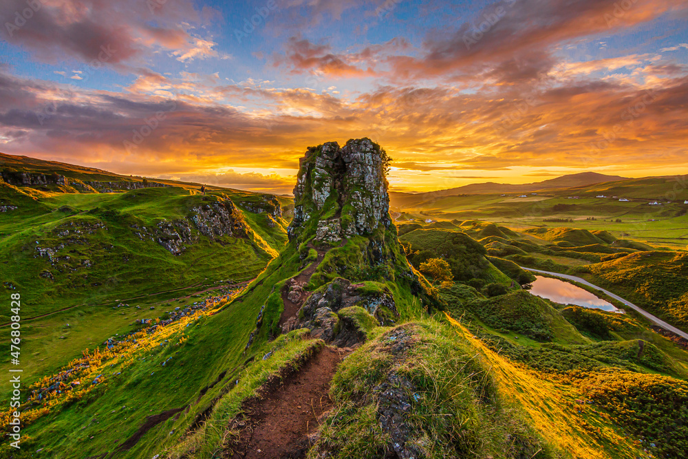 Sunset on the Isle of Skye in Scotland. Castle Ewen mountain hill with paths and meadow in the evening. Red and orange clouds in the sky. small lake with road. Sunshine in the valley