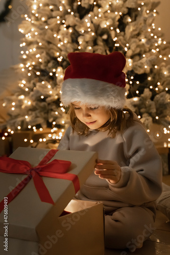Christmas spirit. Happy little smiling girl opening gift box sitting on the floor near the Christmas tree. Happy girl unwrapping christmas present looking in box. Holidays and celebrations concept