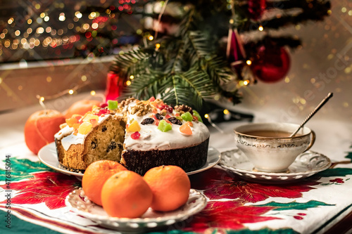 Christmas stollen with glaze, candied fruits and nuts on a saucer with a cup of tea, tangerines near a small Christmas tree. A festively set table for a Christmas Eve tea party on a background of boke