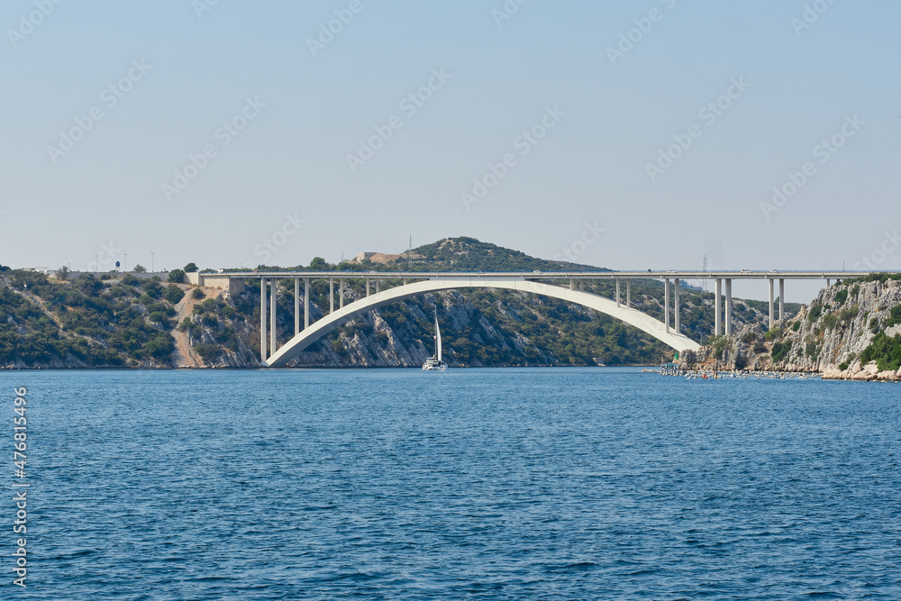 Yachts sail along the gulf of the Adriatic Sea towards the large arched road bridge that connects the islands of Croatia