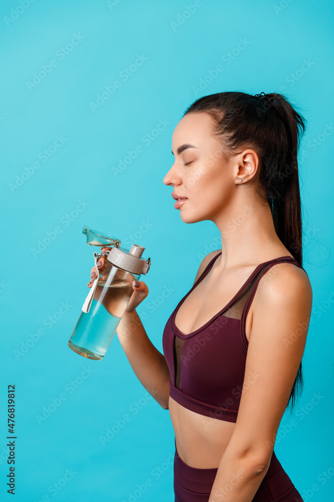beautiful young brunette woman in sportswear drinks water from a bottle on a blue background beautiful young brunette woman in sportswear drinks water from a bottle on a blue background