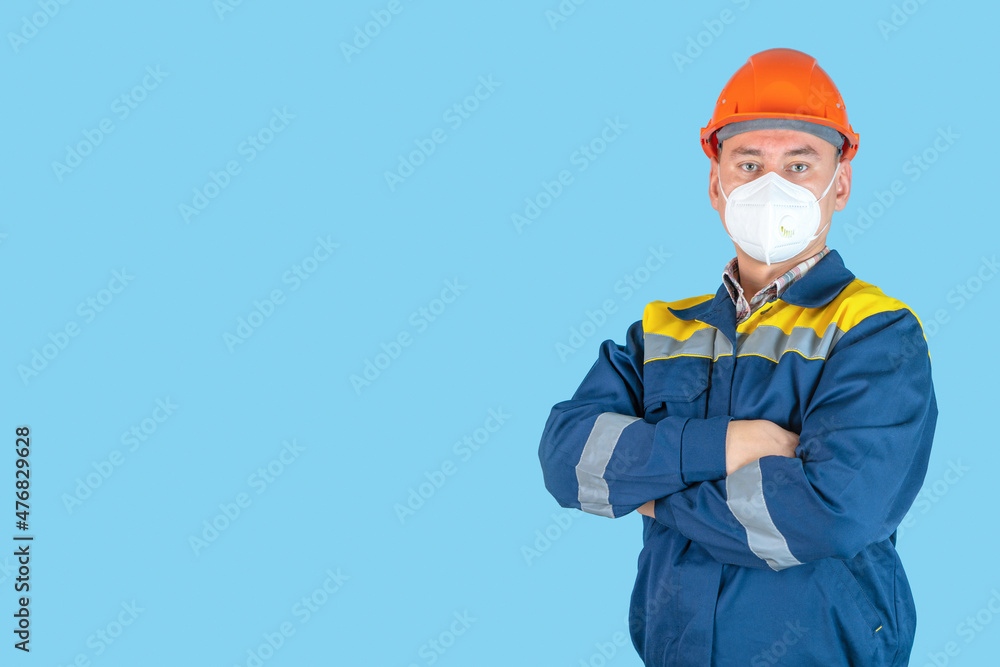 A professional builder in an orange hardhat and a protective mask stands with his arms crossed on a blue background. Copyspace A professional builder in an orange hardhat and a protective mask stands with his arms crossed on a blue background. Copyspace