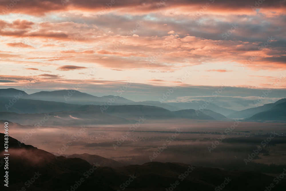 Surreal mountain landscape with pink low clouds above village among mountains silhouettes under dawn cloudy sky. Atmospheric alpine scenery of countryside in low clouds in sundown magenta color.