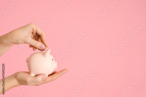 A woman's hand holds a piggy bank in the palm of her hand on a pink background and puts a coin in it, copyspace