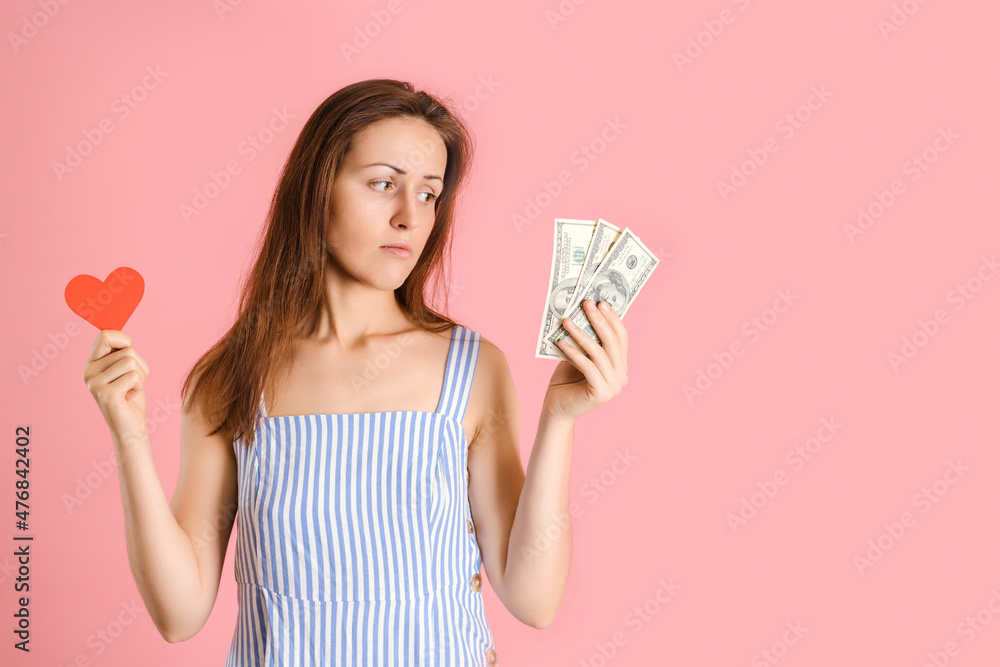 The choice between love and money. A woman holds a heart symbol in her hands and looks at the money in her hand. Studio shot on a pink background The choice between love and money. A woman holds a heart symbol in her hands and looks at the money in her hand. Studio shot on a pink background