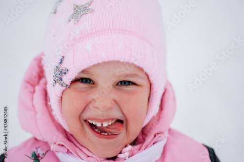 Closeup photo of female kid smiling and sticking tongue out of mouth wearing warm winter clothes in forest. Astonishing background full of white color and snow. 