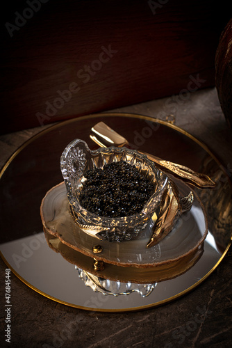 Black caviar in a crystal bowl, decorated with golden sturgeon on a dark concrete background.