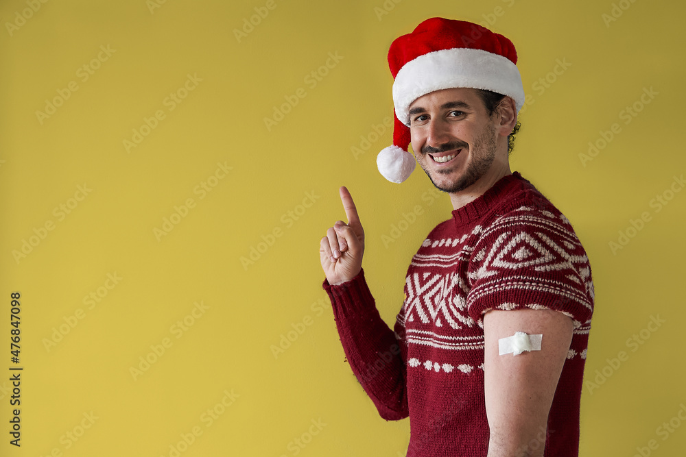 Christmas vaccination campaing. Smiling young man with santa hat and red sweater with bandage in arm points away over gold yellow background Christmas vaccination campaing. Smiling young man with santa hat and red sweater with bandage in arm points away over gold yellow background