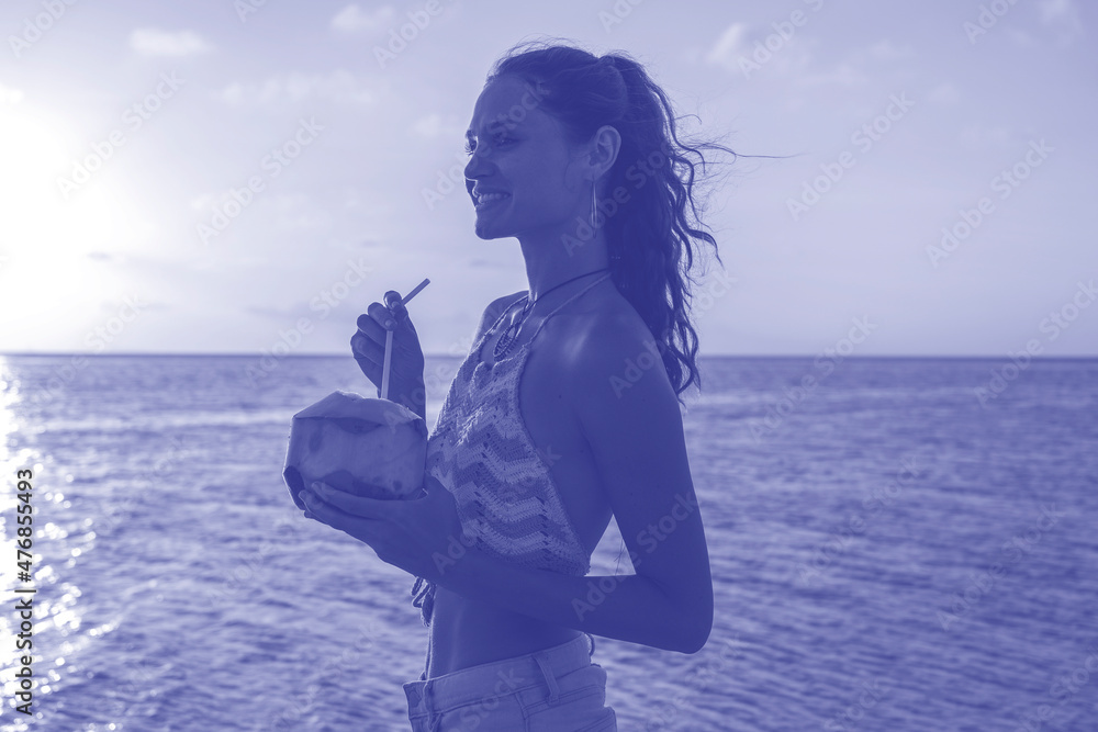 Young beauty girl drinking coconut at tropical beach near sea water at paradise island at sunset. Trendy color of the year 2022 . Ultra Violet creative and moody color of the picture