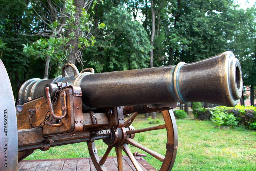 Close-up of an old bronze cannon for throwing balls against a background of green trees. Cannon on wooden wheels. View in isometry. Soft daylight. Summer.