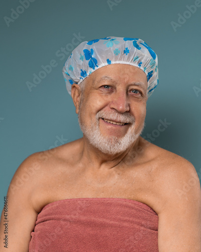 A man with a gray beard aged in a shower hat and a towel, looks and smiling on a blue background, vertical format