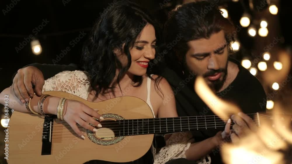 Romantic couple. Brutal guy teaches to play the guitar beloved girl in white dress. They sit in an embrace in the night forest by the bonfire on the grass. Garlands in the background. Romantic couple. Brutal guy teaches to play the guitar beloved girl in white dress. They sit in an embrace in the night forest by the bonfire on the grass. Garlands in the background.
