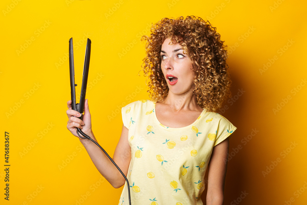 young woman with curly hair looks in surprise at the hair straightener on a yellow background young woman with curly hair looks in surprise at the hair straightener on a yellow background