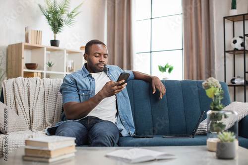 Handsome african man in casual wear talking on smartphone while resting on comfy couch. Young guy having mobile conversation during free time at home.