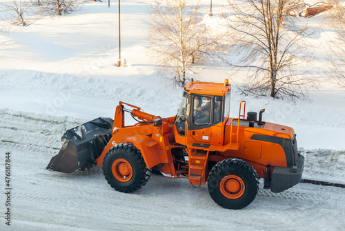 Big orange tractor cleans up snow from the road and loads it into the truck. Cleaning and cleaning of roads in the city from snow in winter