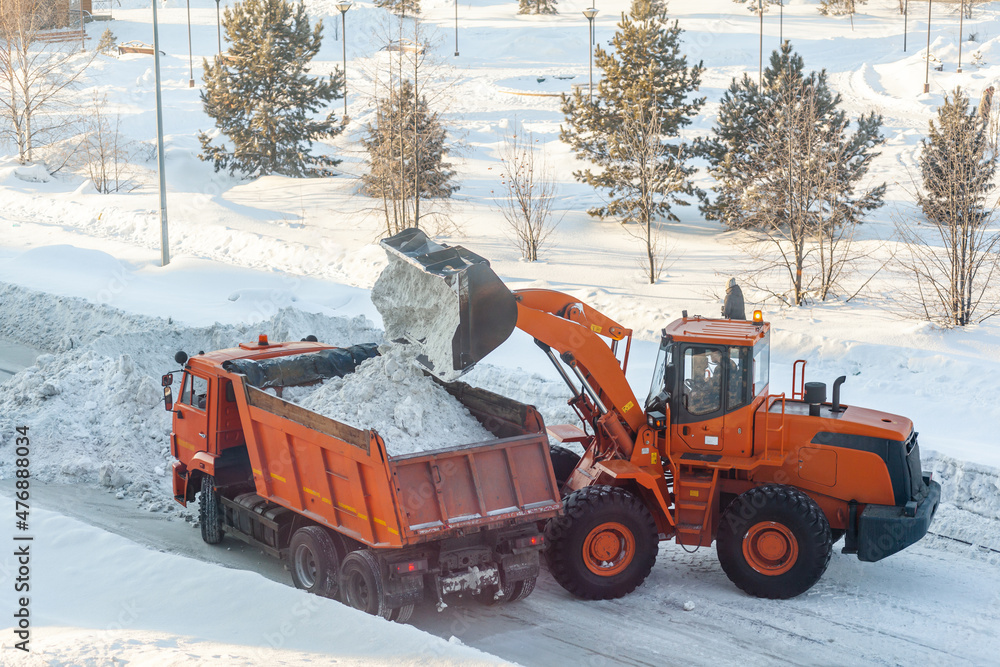 Big orange tractor cleans up snow from the road and loads it into the truck. Cleaning and cleaning of roads in the city from snow in winter Big orange tractor cleans up snow from the road and loads it into the truck. Cleaning and cleaning of roads in the city from snow in winter