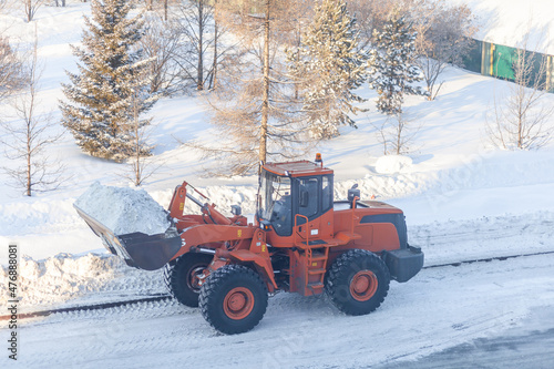 Big orange tractor cleans up snow from the road and loads it into the truck. Cleaning and cleaning of roads in the city from snow in winter