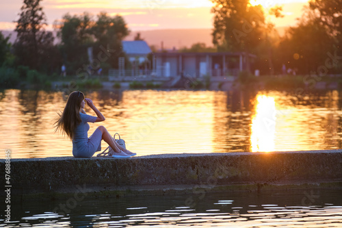 Side view of lonely woman sitting alone on lake shore on warm evening. Solitude and relaxing in nature concept