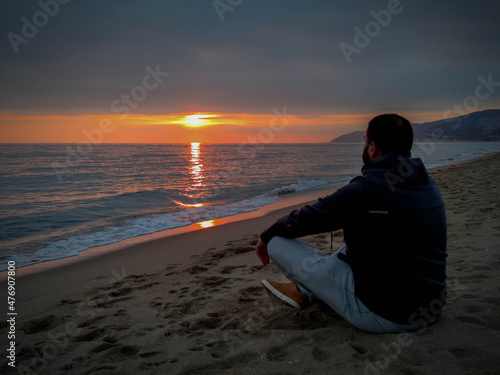 Young man sitting on the beach regarding beautiful sunset over mediterranean sea at autumn 