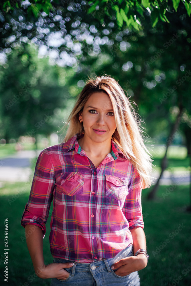 Portrait photo of adult shining blonde woman with hands in pockets in park wearing checkered shirt and jeans skirt in daytime. Adorable background full of green.