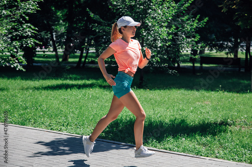running smiling young sporty woman with watch in park in pink t-shirt, green shorts, white cap and trainers on background of green trees and lawn on grey tiles in sunny summer day from the front.