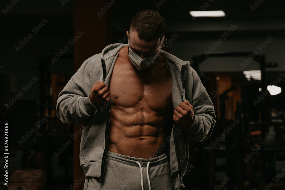 A bodybuilder in a face mask is opening his hoodie to demonstrate his physique. A bodybuilder in a face mask is opening his hoodie to demonstrate his physique.