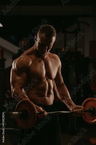 A bodybuilder with a beard is doing bicep curls with a short barbell in a gym.