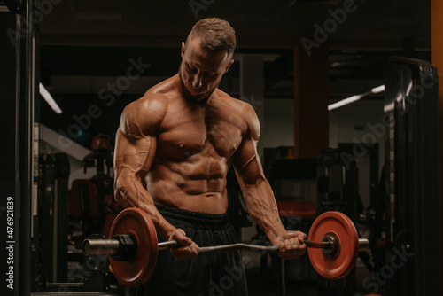 A bodybuilder with a beard is doing bicep curls with a short barbell in a gym.