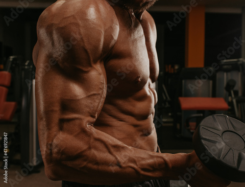 A close up shot of a torso of a bodybuilder who is doing bicep curls in a gym.