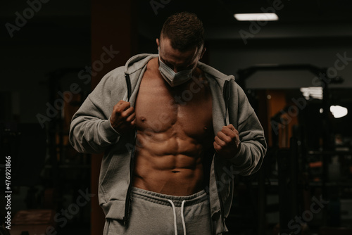 A bodybuilder in a face mask is opening his hoodie to demonstrate his physique.