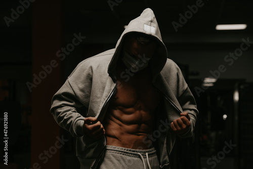 Bodybuilder in a hood and a face mask is opening a hoodie to demonstrate his abs