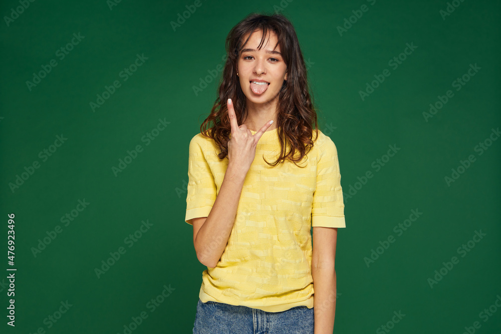 Modern cool young girl show rock-n-roll gesture, stick tongue, female do devil horns hand sign on green background Modern cool young girl show rock-n-roll gesture, stick tongue, female do devil horns hand sign on green background