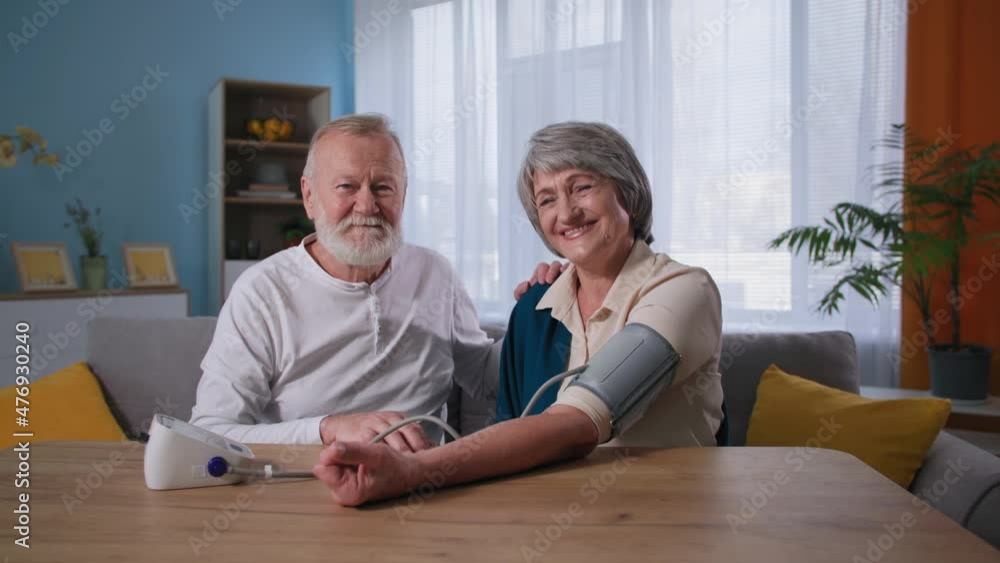 elderly happy couple looking at camera while taking blood pressure, husband using blood pressure monitor for wife with hypertension at home elderly happy couple looking at camera while taking blood pressure, husband using blood pressure monitor for wife with hypertension at home