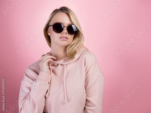 beautiful young woman with blonde hair wearing dark sunglasses. In a pink hoodie on a pink background, studio portrait