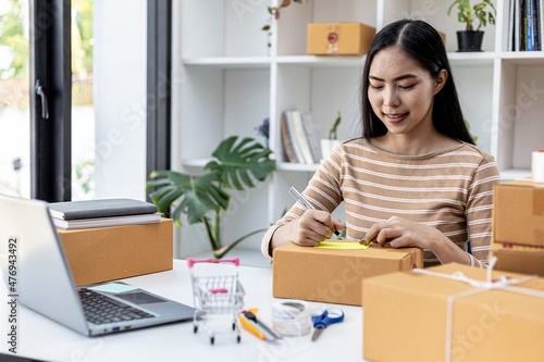 Asian woman writing customer's shipping information on parcel box, she owns an online store, she ships products to customers through a private courier company. Online selling concept. © kamiphotos