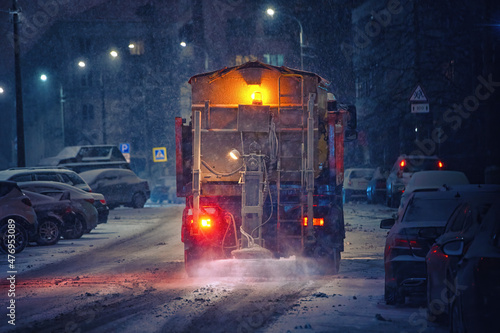 Salt spreading. Snow plow service truck removing snow and spreading salt on snowy city road during blizzard, night work road maintenance. Truck spreading de-icing salt on snowy and icy asphalt road