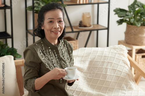 Smiling elegant senior woman with cup of tea sitting on sofa in living room