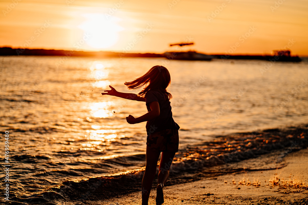 at sunset. kid girl walks along seashore, collects stones and throws into water